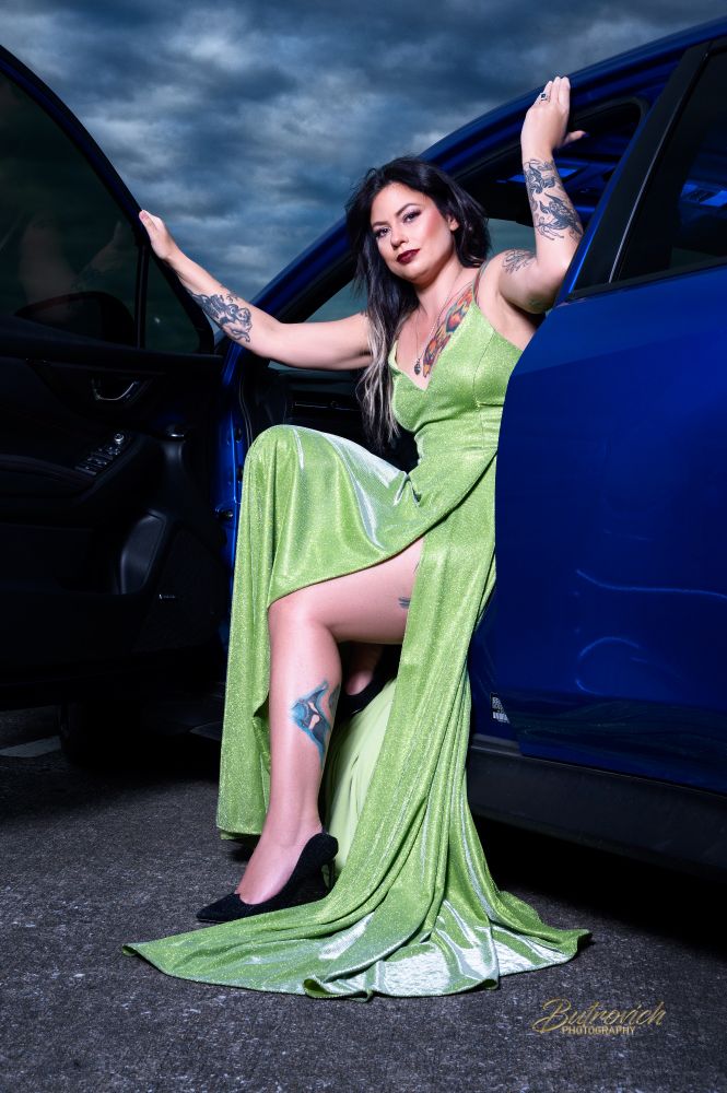 This powerful image showcases a confident, tattooed woman stepping out of a sleek blue car, wearing a bold, shimmering lime-green gown. The moody, dramatic sky and sharp contrast lighting evoke strength, rebellion, and glamor. Her pose—arms extended across both doors—exudes dominance and freedom, while the car serves as a symbol of independence and luxury. It’s a perfect fusion of elegance, edge, and empowerment.