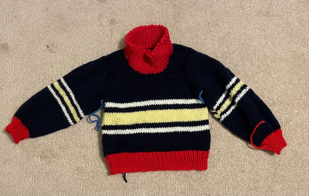 Toddler size sweater with navy body, red ribbed neck, cuffs, and waist, and yellow and white stripes. Loose yarns, as it needs finishing 