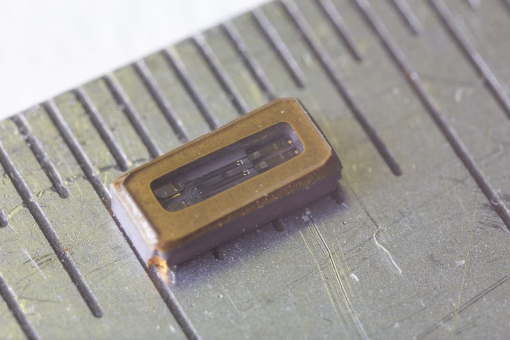Macro photo of a ruler with surface mount 32.768kHz crystal component, showing about 3mm in length. The glass top of the components shows the internals