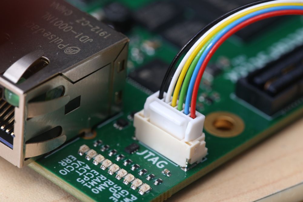 Circuit board with a connector labelled JTAG. The circuit board has LEDs along the front with various silkscreen labels. The connector has cables attached that are vibrant colours. Red, blue, green, yellow, white and black.