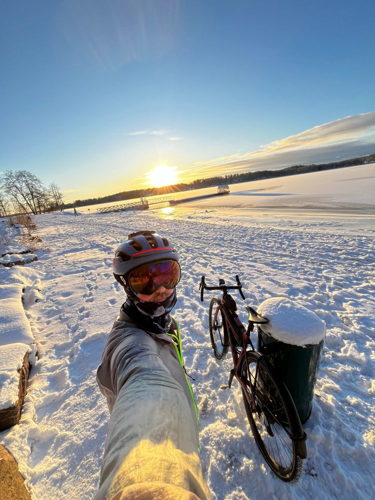 Selfie of a bike rider with helmet and goggles in a wintry scene on a frozen shore with bright sunshine. 