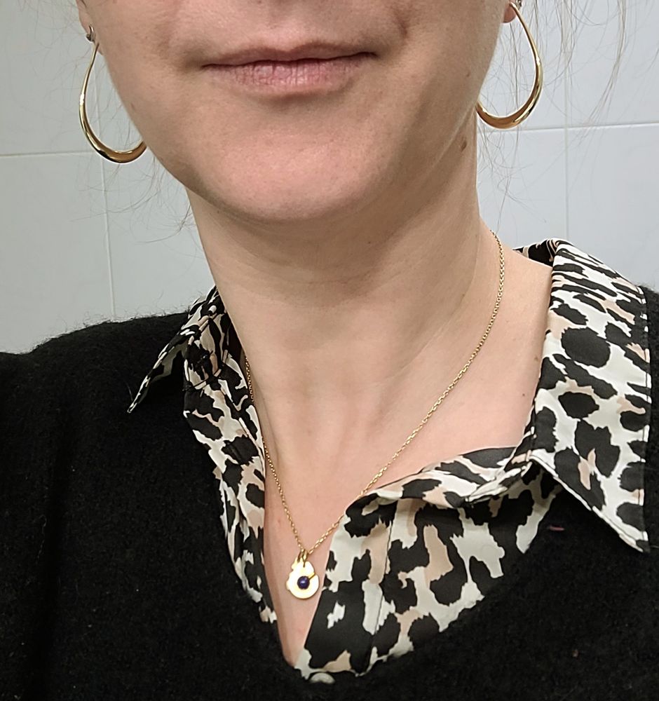 Photo de moi, de près. On voit le bas de mon visage et ma gorge. Aux oreilles, e porte des créoles en forme de goutte, dorées. En vêtement, je porte un pull col V noir duquel sort le col d'une chemise à motif léopard, et une chaîne dorée avec un petit pendentif doré à forme de menotte avec une perle de Lapis Lazuli au centre. 