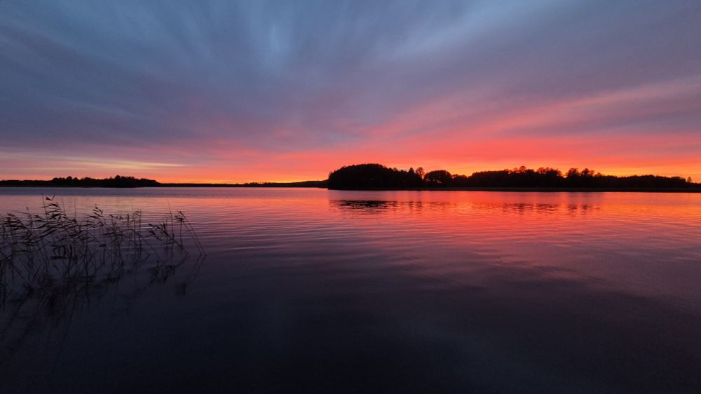 Iltarusko kuvattuna järven rannalta.
Taivas keltainen, oranssinen, punainen, vaaleanpunainen, siniharmaa...
Keskellä saaria, jotka heijastuvat taivaan tavoin osin veteen. Melko tyyntä. Vasemmalla alhaalla kaisloja.