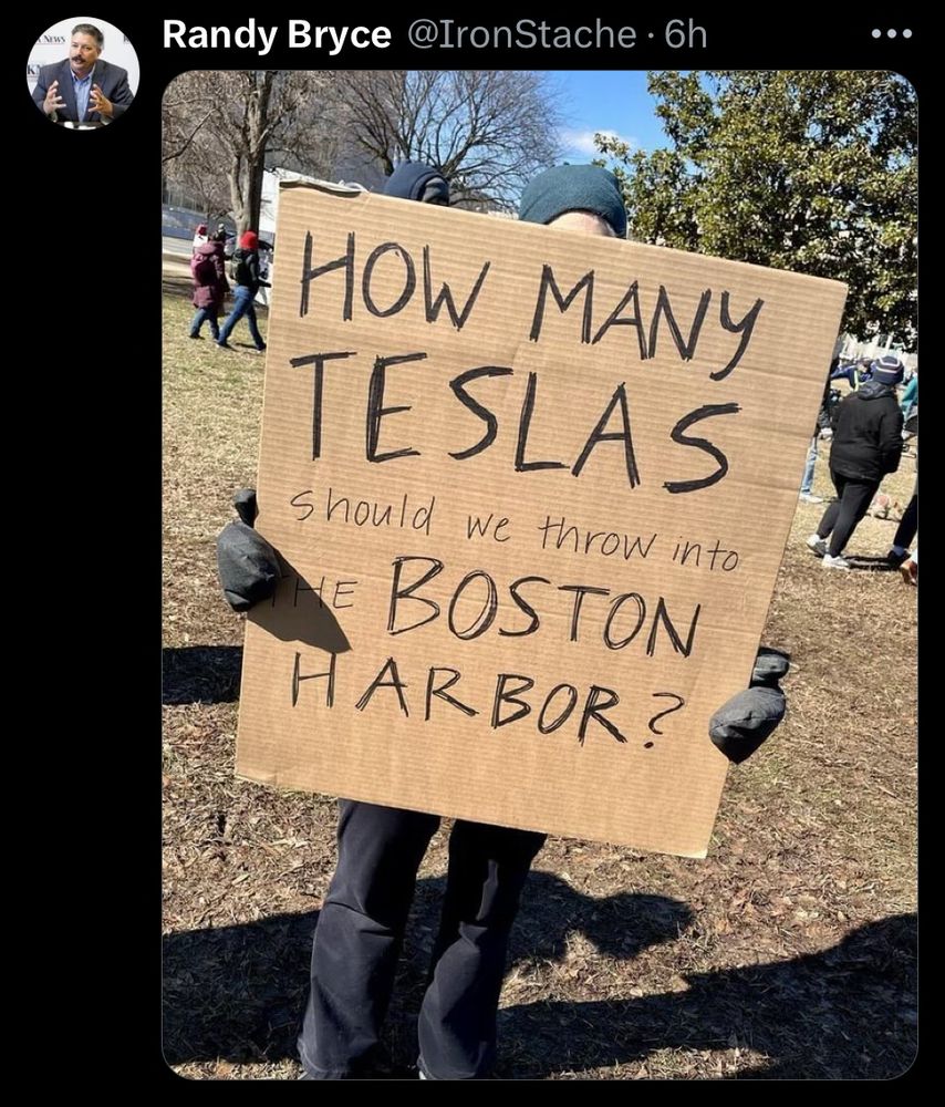 Protester holding a cardboard sign that reads, “How many Teslas should we throw into the Boston harbor?”