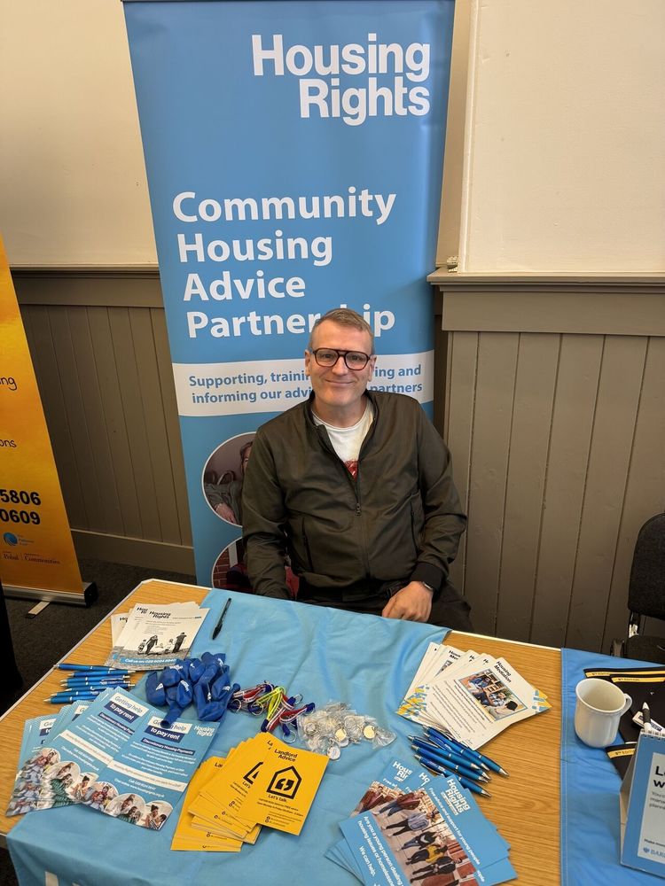 Jonathan is wearing glasses and a green jacket. He is sitting at a table with Housing Rights merchandise. Behind him is a pop up with white text "Community Housing Advice Partnership".