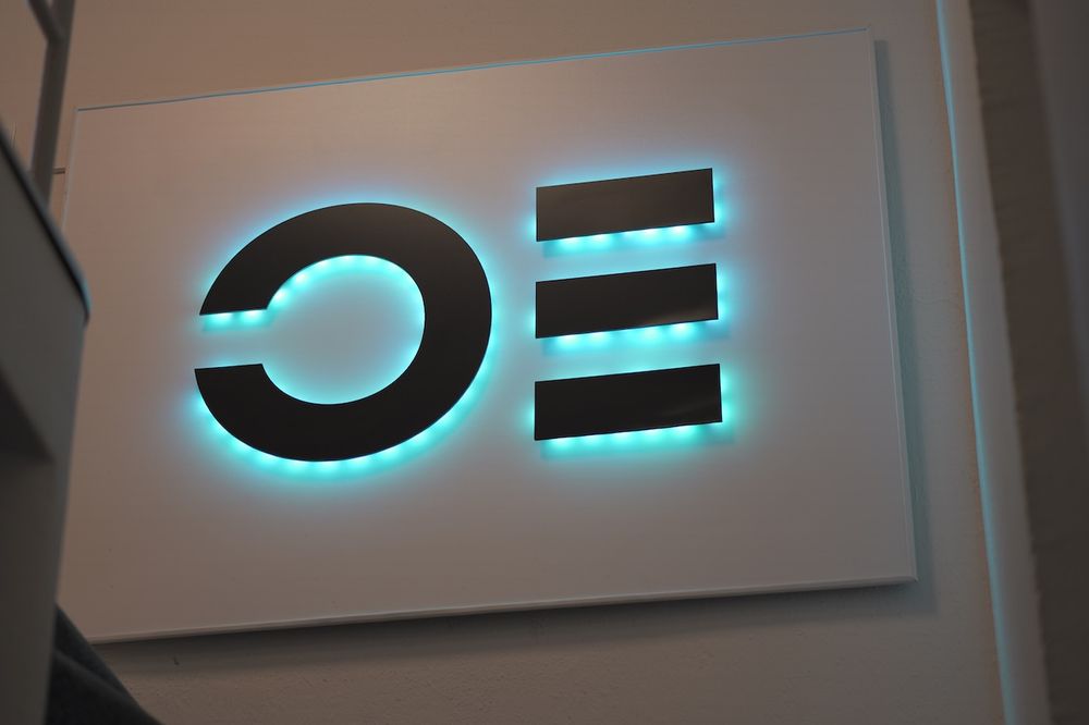 Ops One Logo-Installation auf der Treppe in unserem Büro: Erhobenes schwarzes Logo auf weissem Grund, mit grünen LED hinterleuchtet.