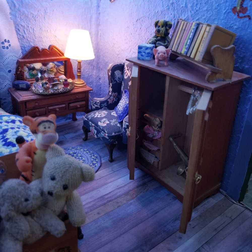 Vanity with perfume tray and lamp sits next to a comfy blue flowered chair. The wardrobe has drawers filled with clothes and a saxophone is on the other side. Stuffed animals and a Tigger are in a trunk at the end of the bed.