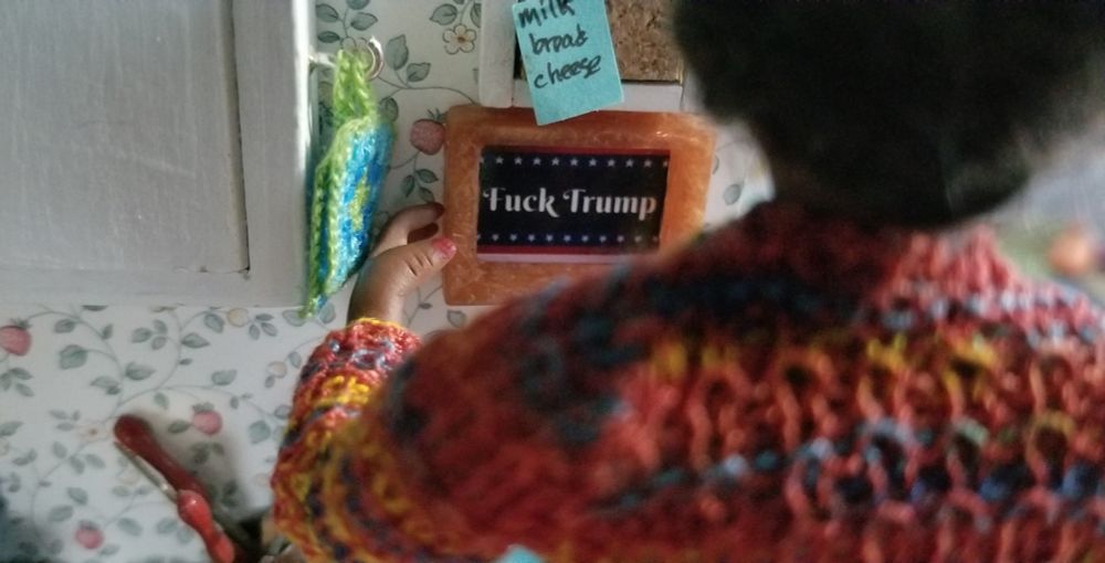 Miniature scene: Doll with back to camera adjusting framed print saying fuck trump under a bulletin board with a grocery list of milk, bread and cheese. Blue and green crochet potholders hang from an upper cabinet. 