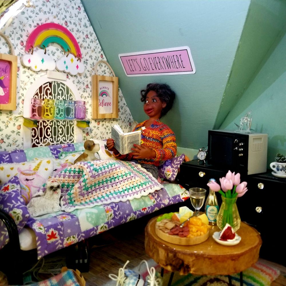 Trixie in her efficiency apartment on her quilt covered futon with her granny square blanket and dogs to keep her warm and comfy. On the round wood coffee table is a cheese tray, glass and bottle of wine, a vase of tulips, and a piece of cake. A sign over her head says Let's Go Everywhere.