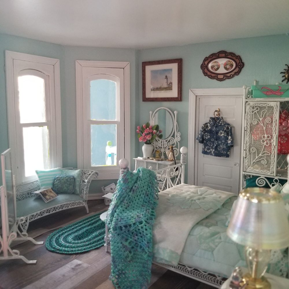 This view of the room shows a white standing mirror and a chaise with aqua cushions and pillows and an open book. There is a vanity with a vase of flowers and some bottles of perfume. Next to the vanity is the door with a Hawaiian shirt on the back and a wardrobe with another shirt inside. Paintings hang on the wall.
