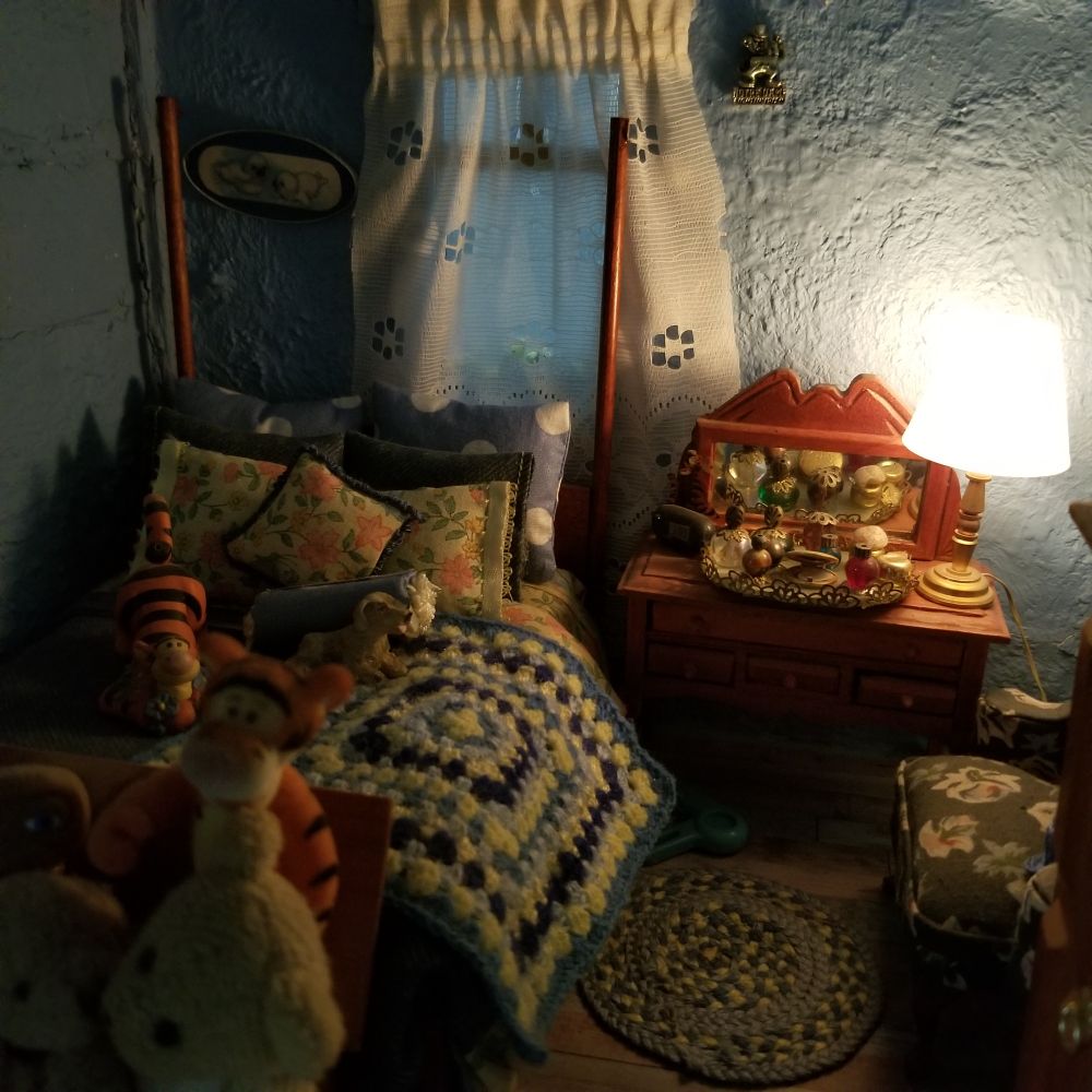 Miniature bedroom includes a 4 poster bed beneath a white curtained window; the bed dressed in blue and yellow flowered bedding and crochet blanket. There are stuffed Tiggers and a dog on the bed. A vanity table beside bed holds tray of perfumes and a lamp to read by while sitting in the blue flowered chair.