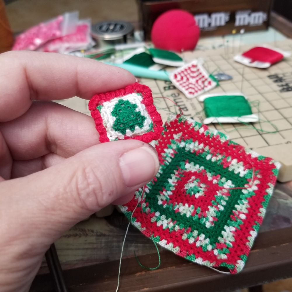 Hand holding ring crochet pillow with a green tree and a red ruffle. Behind is a WIP of a granny square blanket in holiday colors. Behind that is a WIP of a red and white pillow getting a green ruffle; green and red embroidery floss. And behind that is my clown nose because why not?