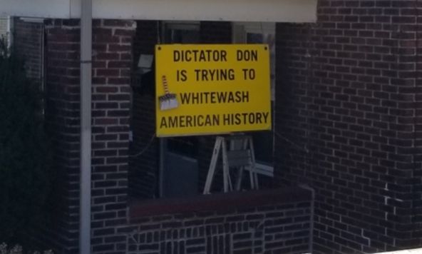 Brick porch with a yellow sign with black lettering saying "DICTATOR DON IS TRYING TO WHITEWASH AMERICAN HISTORY" with a paintbrush filled with white paint. 