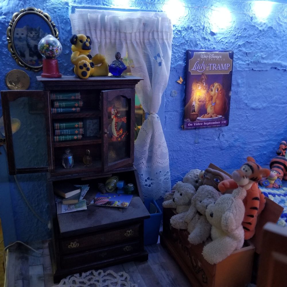 Desk with hutch filled with books, on top sits a gumball machine, a yellow bear and a glass perfume bottle. Behind is a white eyelet curtained window. At the end of the bed is a trunk filled with stuffed animals.