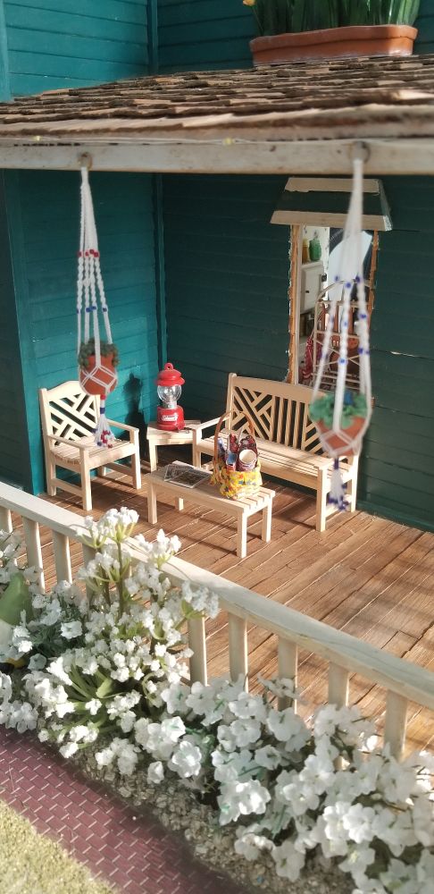 On the front porch is a wooden set of couch and chair to relax upon. White flowering shrubs are along the outside of the porch. Hanging macrame plant hangers with plants in them complete the look.