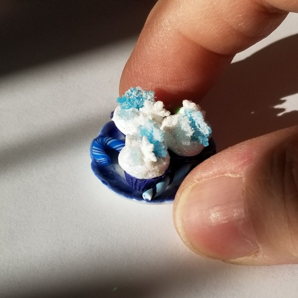 Two fingers holding a miniature plate in the sunshine. On the plate are 3 cupcakes frosted in white with blue and white snowflakes on top and blue and white candy canes. 