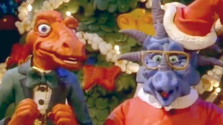 Rex, an orange t-rex in a tuxedo, and Herb, a purple ??? with lots of horns in a Santa suit, standing in front of a Christmas tree