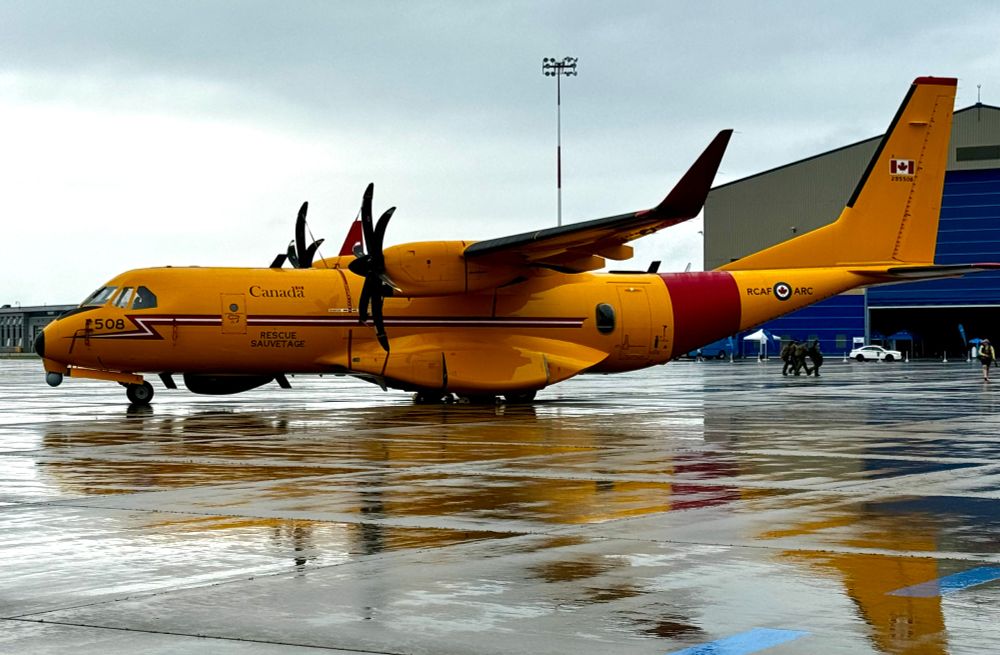 A bright yellow mid-sized cargo airplane in RCAF search and rescue markings on a wet parking area with grey skies overhead.