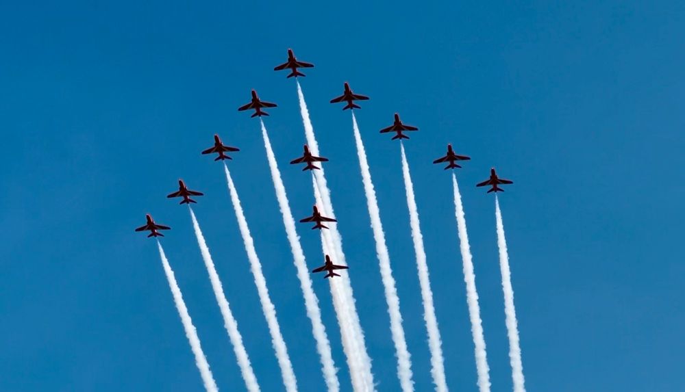 12 RAF Red Arrows jets, trailing decorative white smoke, flying overhead in formation. 