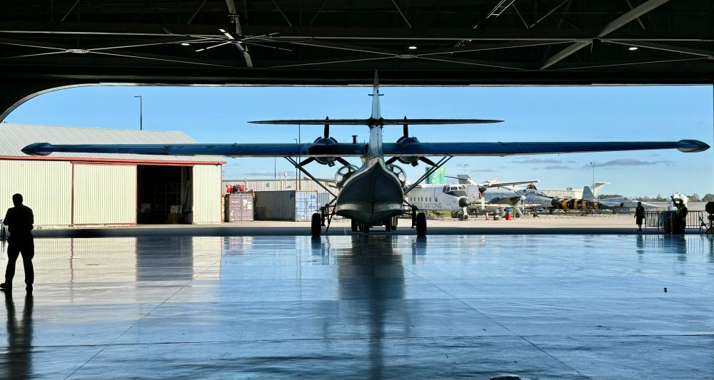 It is a WW2 vintage PBY-5 Canso flying boat with its reflection on a shiny floor as it is pulled out of a hangar on a sunny day with a blue sky in the background.