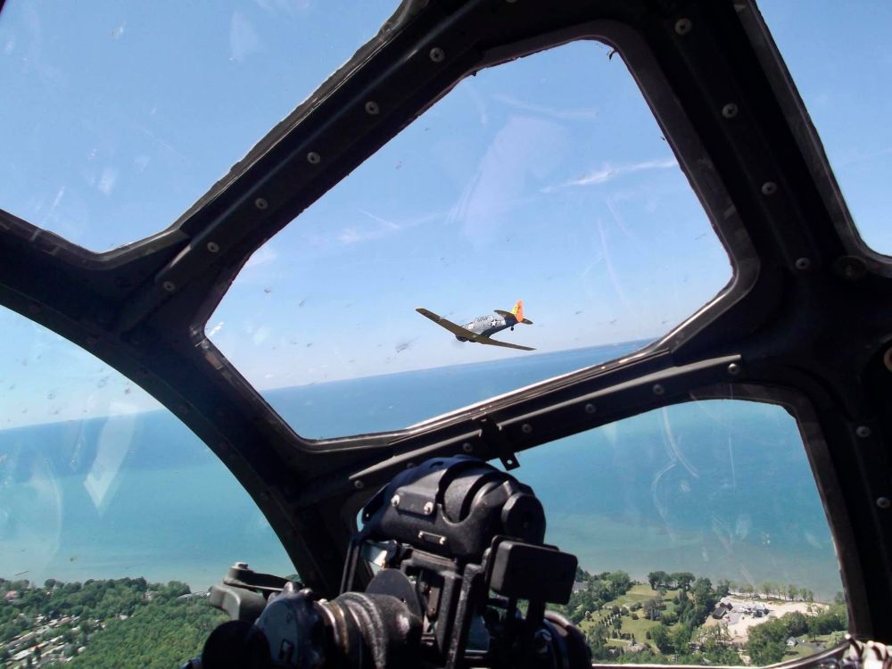 Seeing a US Navy SNJ training airplane flying over a shoreline while looking through one of the forward windows of a B-29 Superfortress while sitting in the bombardier's seat at the very front of the cockpit.