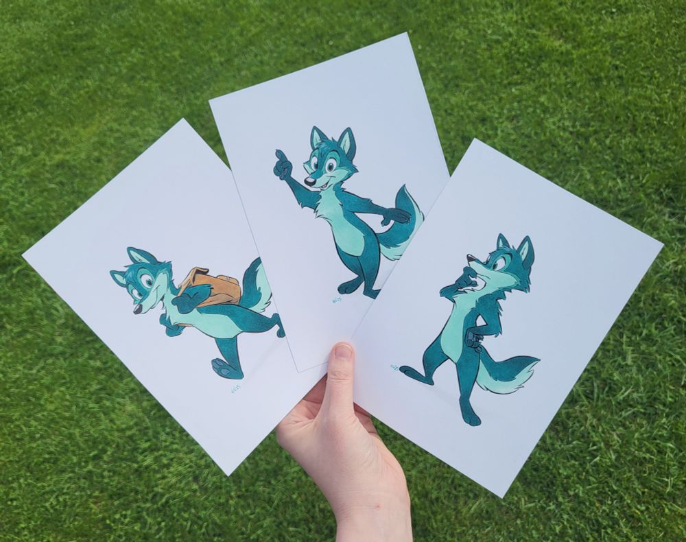 My hand holding up three A5 sheets of bristol, with on it three different traditional drawings of Greeny, a green cartoon wolf. The drawings are inked with india ink and colored with alcohol markers.
