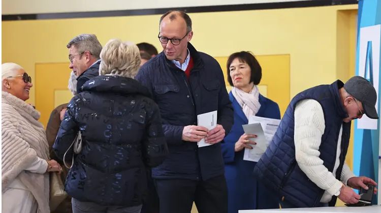 Friedrich Merz, un señor de mediana edad, alto, medio calvo y con gafas metálicas negras, un chaquetón azul marino, rebeca de señor mayor y camisa azul, espera para votar en su colegio. 