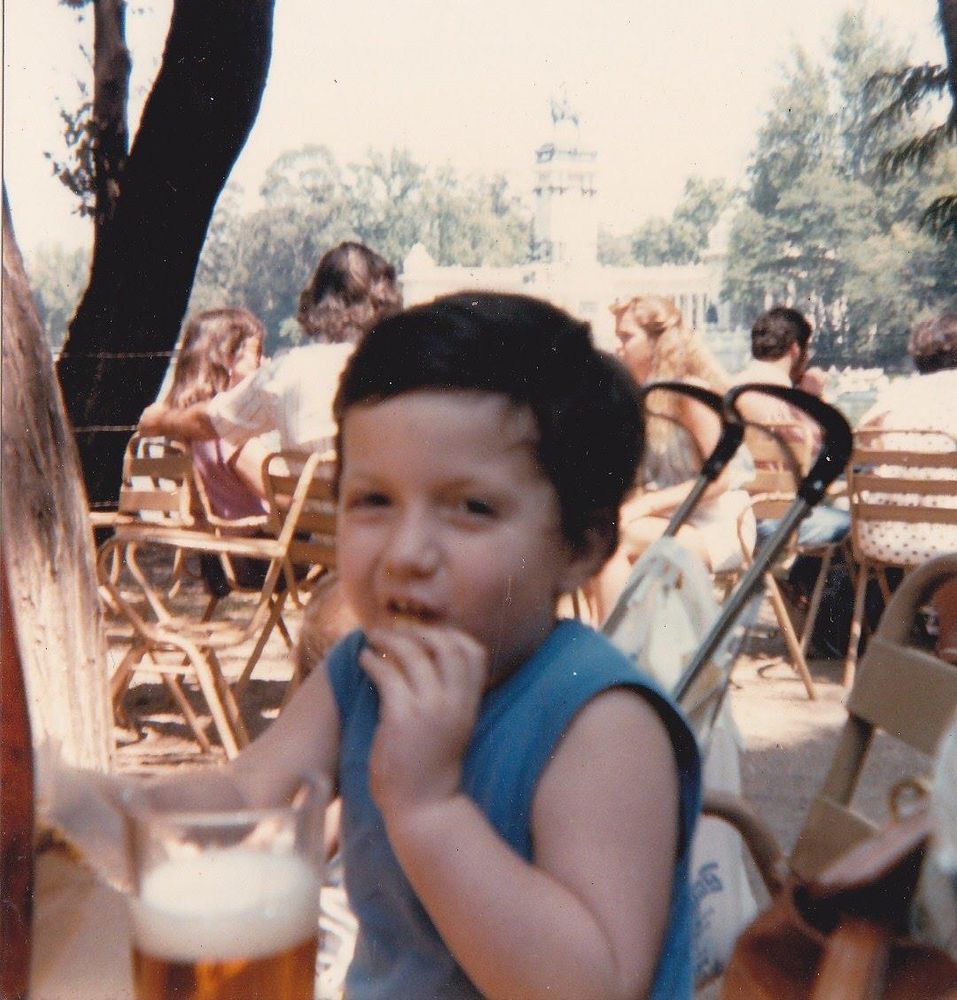 Pequeño Thiago Ferrer, de camiseta azul sin mangas, comiendo (probablemente una galleta) en una terraza junto al estanque del Retiro. El carrito de mi hermano está detrás. 