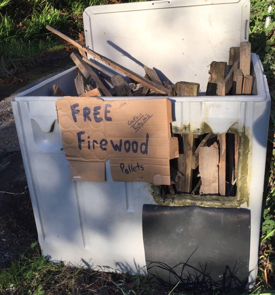 A bin filled with old cut-up pallets. An attached cardboard sign says, “Free firewood—Pallets—caution rusty nails.”