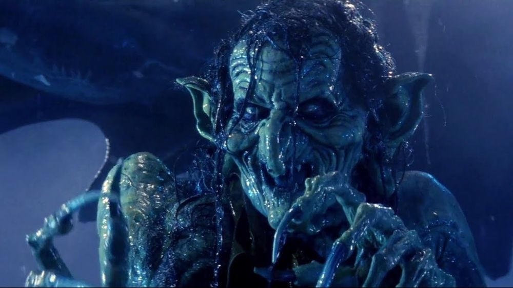 An image of Meg Mucklebones from the movie Legend. She is a slimy, green skinned hag, smiling as she points with one clawed hand. 