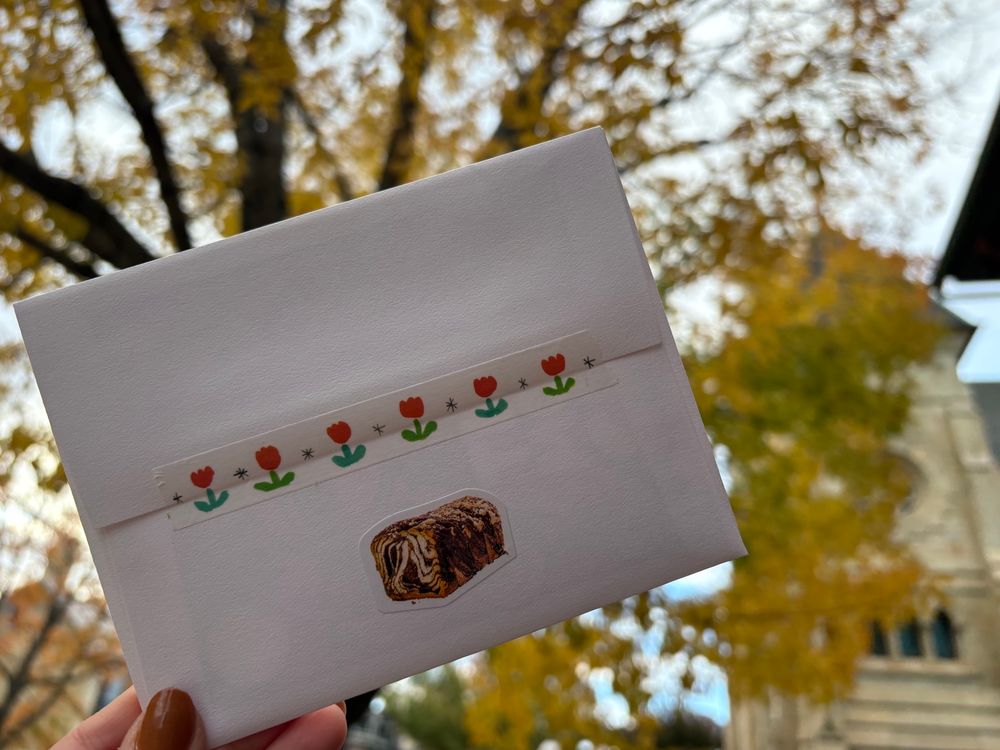 Holding a sealed envelope in front of a tree with yellow leaves; the seal of the envelope has tulip washi tape on it and there is a sticker depicting a beautiful chocolate babka affixed underneath the seal line 