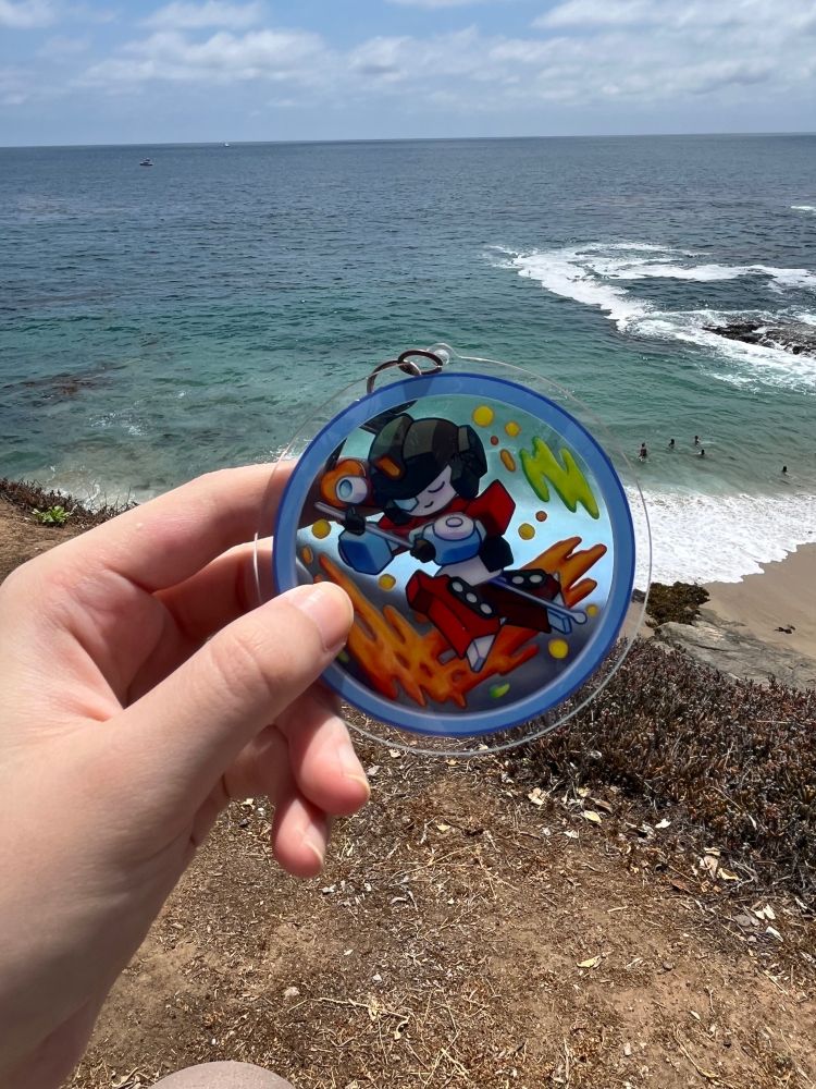 Photograph of a hand holding up an acrylic charm of Perceptor at a beach.