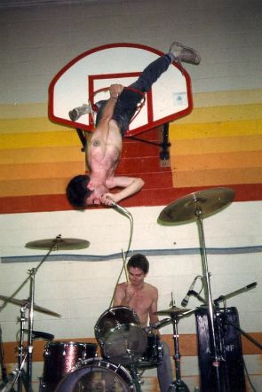 Guy Picciotto, vocalista da banda Fugazi, cantando de ponta-cabeça dentro do arco de uma cesta de basquete de uma quadra de escola feita de palco. Embaixo dele, está o baterista tocando bateria.