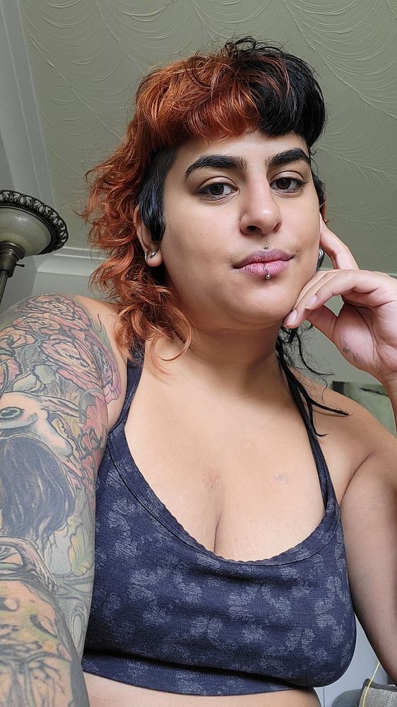 Kitty looks down at the camera, one hand against their cheep and a soft smile. They are a light skinned brown femme with split black and orange hair with shaved sides. They have two lip piercings and a sleeve tattoo. They are wearing a black crop top. 