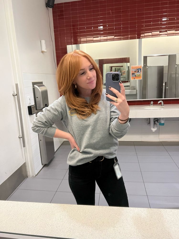 a white woman with redhead wearing a gray, company crew neck sweater and black jeans taking a selfie in the mirror of an office restroom. 