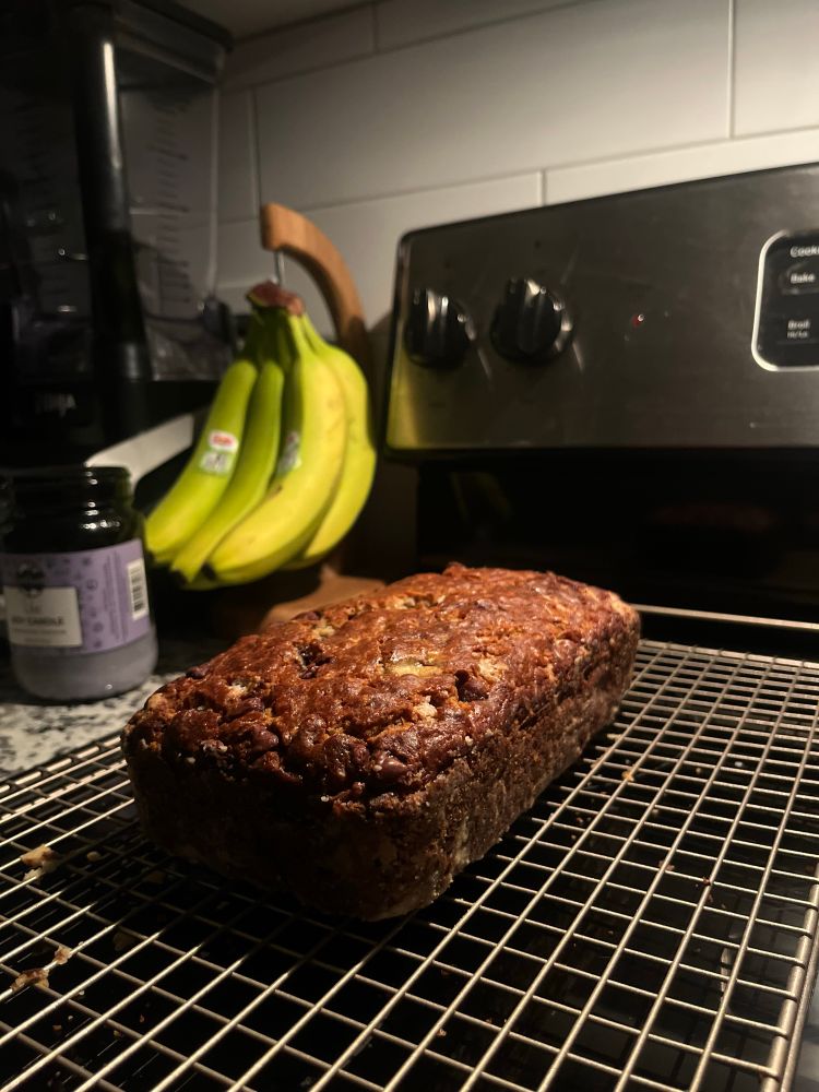 a golden-brown loaf of banana bread cools on a wire rack in a kitchen, with bananas hanging in the background