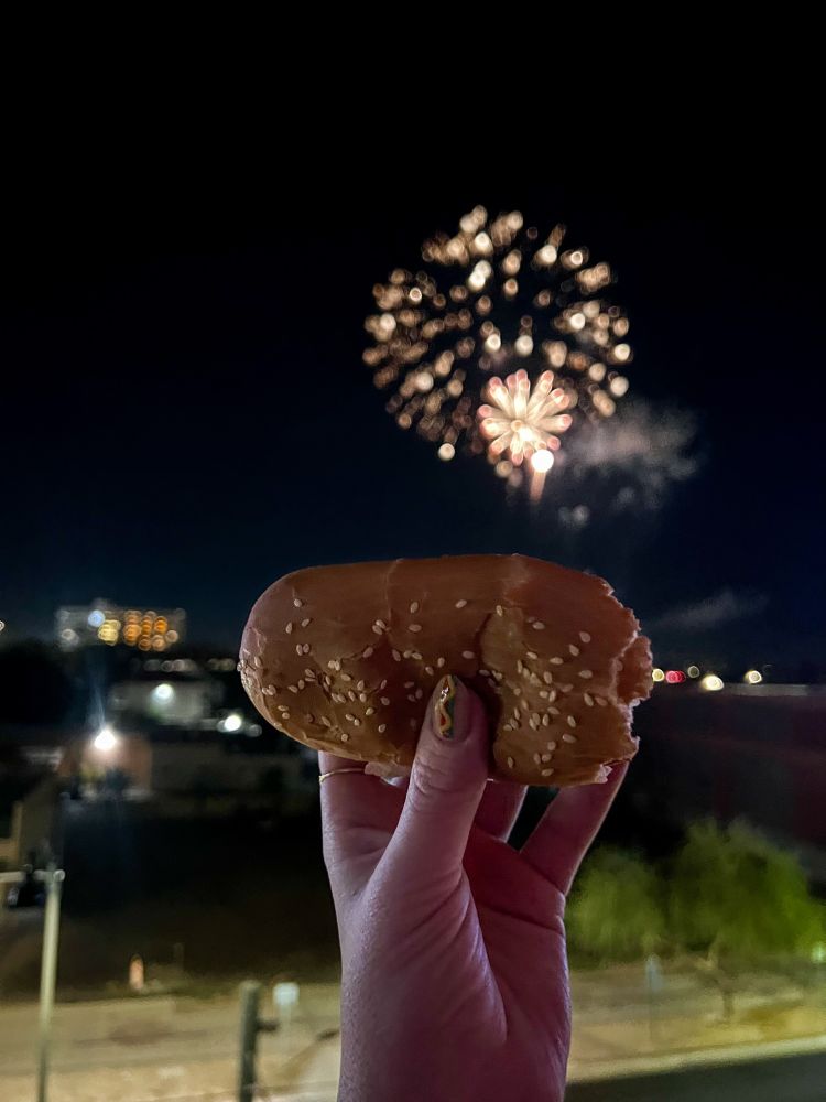 a hand with hot dog nail art holding a hot dog in front of fire works