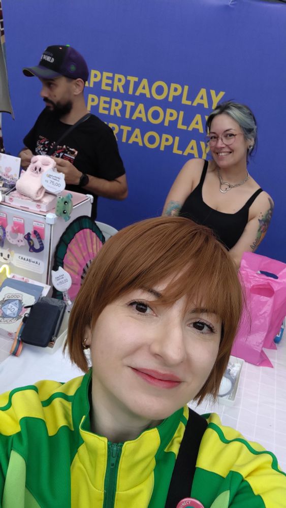Eu, com meu semi-cosplay de Chie de Persona 4, fazendo selfie na frente do estande da Kaol, com ela aparecendo no fundo toda sorridente!