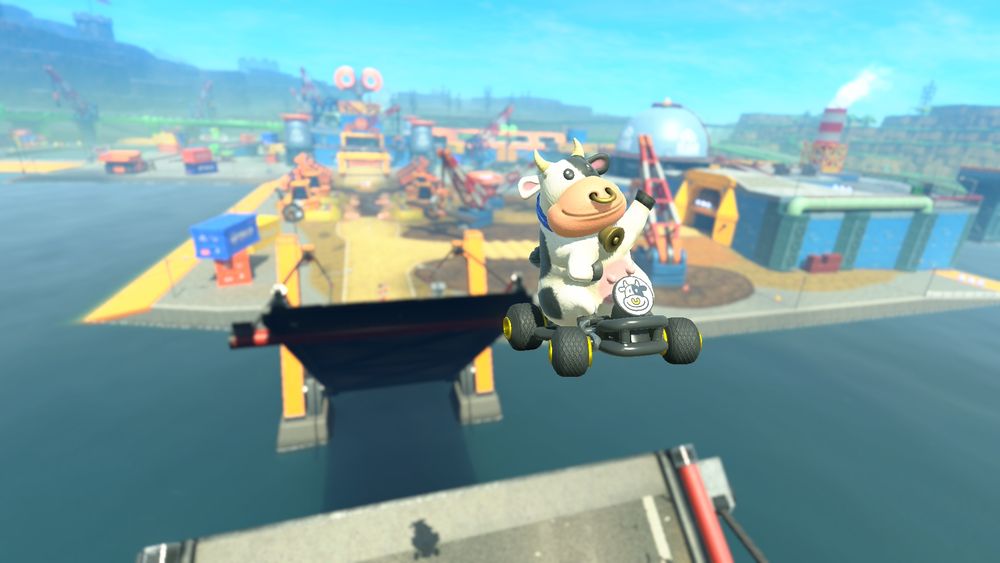 Screenshot of Mario Kart World. A cow is doing a disco pose with one arm in the air after to jumped off a raised drawbridge. Toad Factory can be seen in the background.