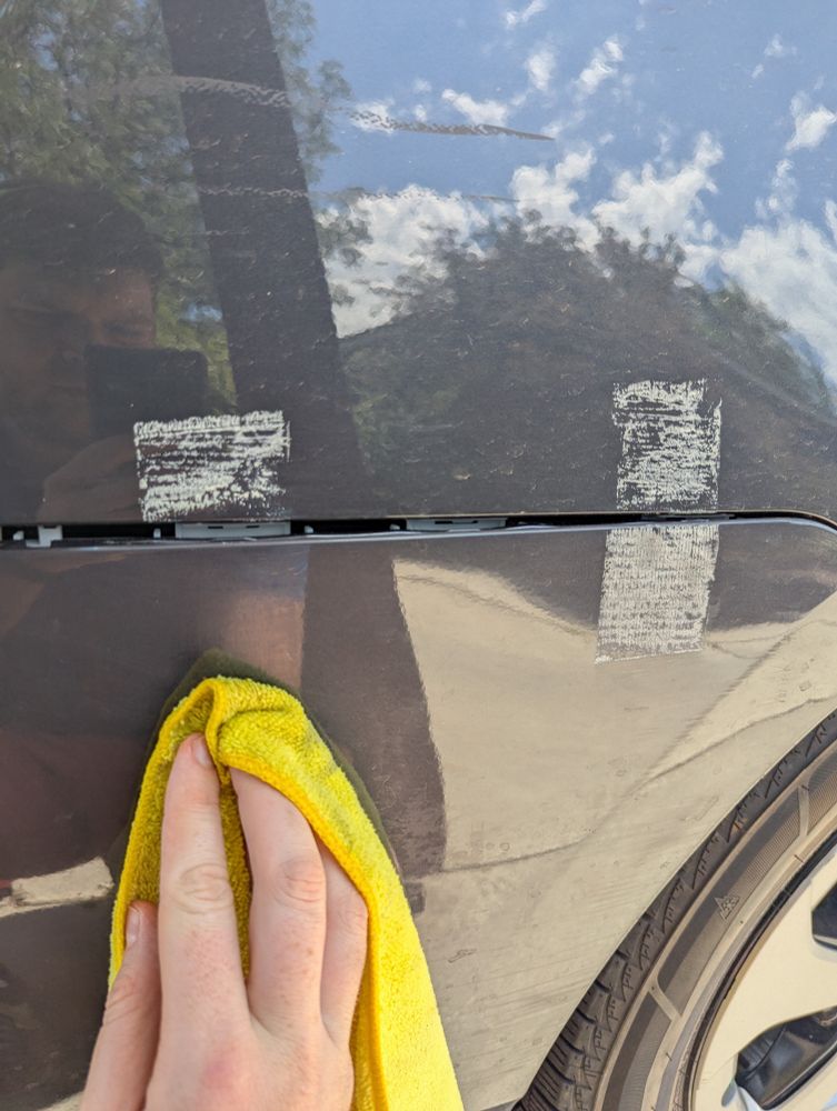 Duct tape residue partially removed from a car exterior and a hand holding a yellow rag covered in gasoline.