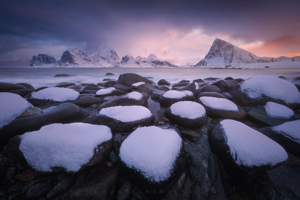 View of a snowy fjord in northern Norway (Lofoten). The foreground is formed by interesting round stones and the background by snowy mountains with a colorful sky.