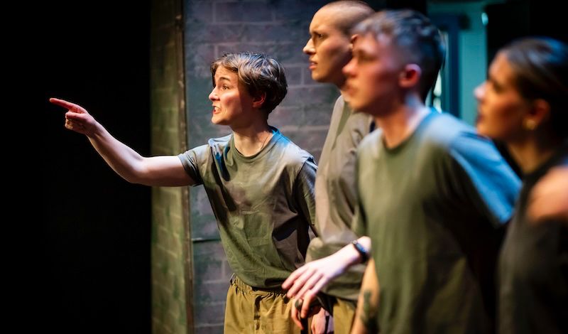 An angry actor as part of a line of characters in fatigues, points angrily in a theatre play.