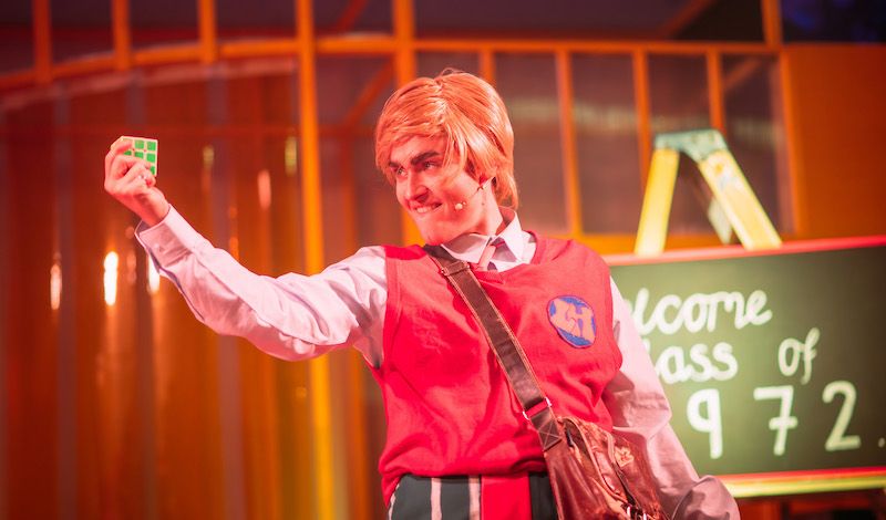 Chloe-Ann Taylor as Johnny McGuffin in a Borris Johnson wig, holds up a Rubik Cube in her right hand.