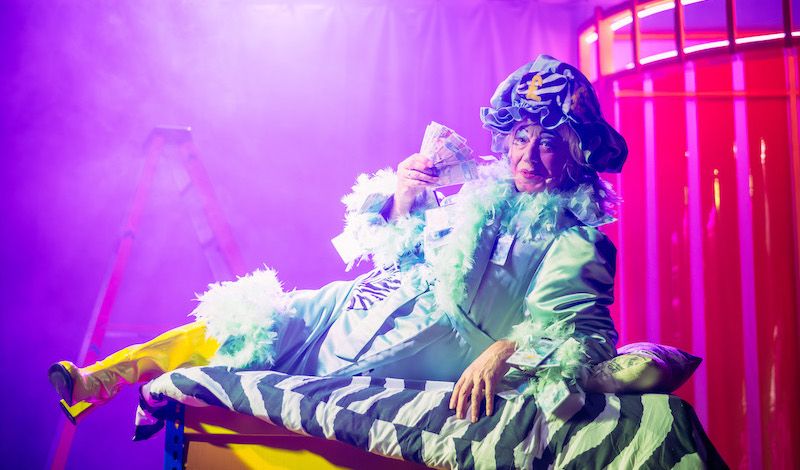 Graham Crammond as Dame Scrooge, reclining on a stripy sofa waving a wadge of cash in her face like a fan.