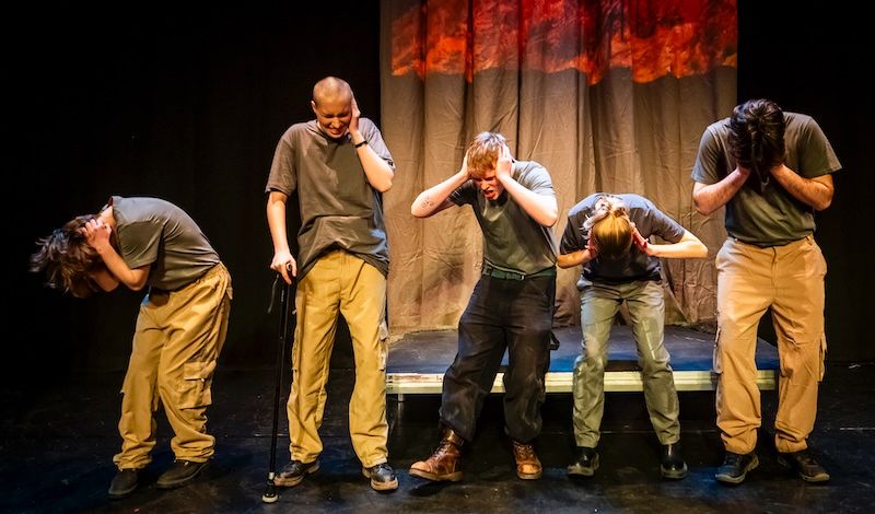 Five young actors dressed in fatigues hold their ears and grimace in a theatre play. The back-cloth shows images of burning.
