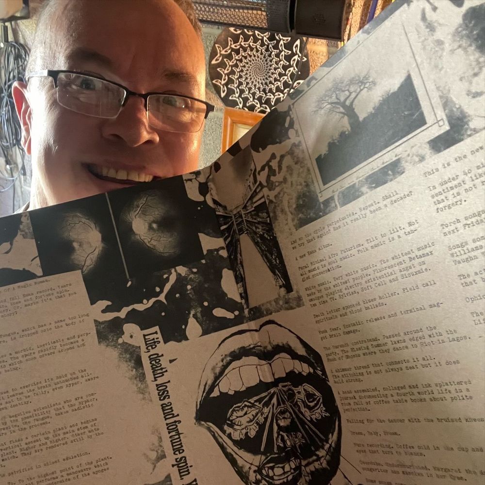 A grinning round-faced white middle aged man holding up the gatefold sleeve of a record to the camera.