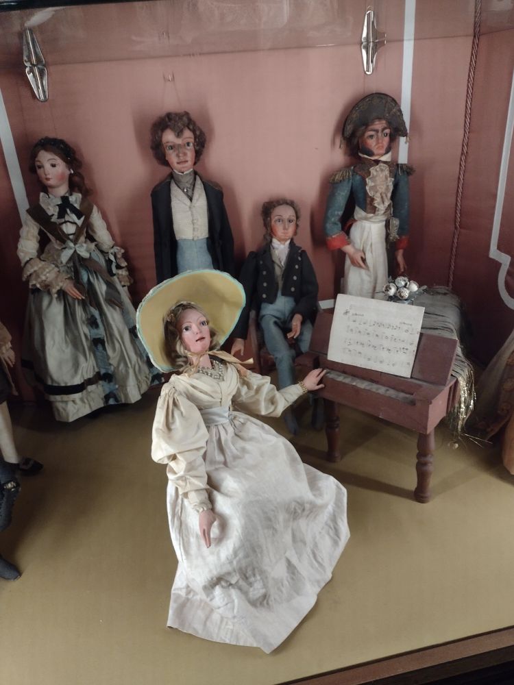 A number of puppets in detailed c19 costume, one of which is seated at a small keyboard instrument 