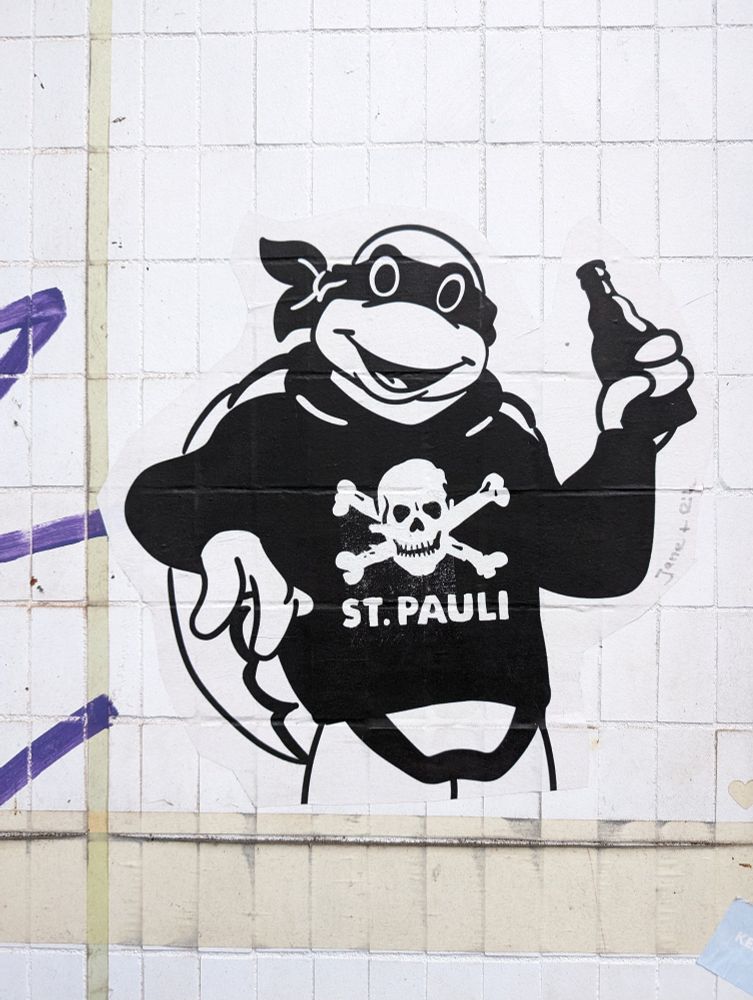 Schwarz-Weiße Grafik, ausgedruckt und an eine Hauswand geklebt:
Oberkörper einer stehenden Comic-Schildkröte (Teenage Mutant Hero Turtles) auf deren Pullover der Totenkopf vom FC Sankt Pauli sowie die Worte "St. Pauli" prangen. Sie hält eine Bierflasche in der linken Hand und lehnt mit dem rechten Ellenbogen auf einem nicht sichtbaren Gegenstand.