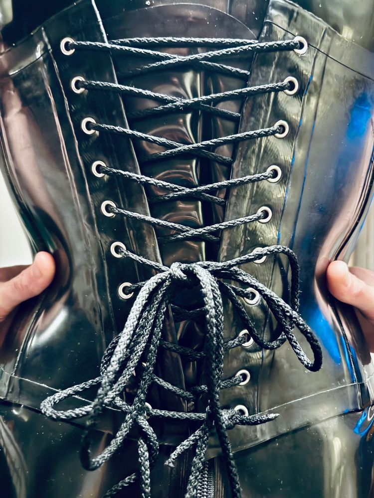 Shot of the back of a black rubber corset that is being worn over a black rubber catsuit. You can see how tight the lacing is 