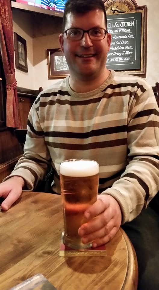 A picture of me from 2015, sitting at a pub table and holding a pint of lager in my left hand; my hair is cropped short, and I'm wearing glasses with blue metallic frames, and a light brown sweater with white, black, and dark brown stripes.